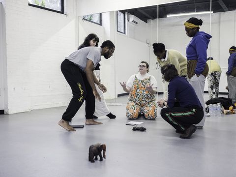 People gather on a dance studio floor, with one person seated and others standing or crouching nearby.