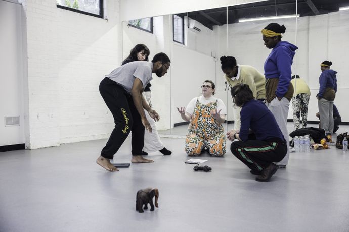 People gather on a dance studio floor, with one person seated and others standing or crouching nearby.