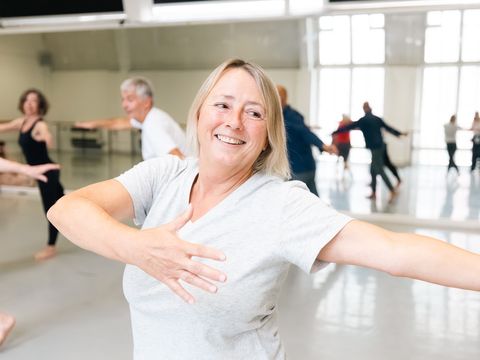 A woman enjoying a dance class, smiling and spreading her arms wide, with other participants visible in a spacious mirrored studio.
