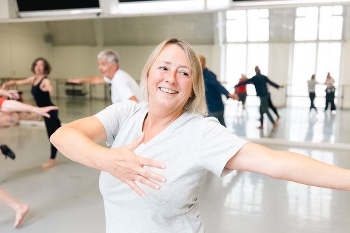 A woman enjoying a dance class, smiling and spreading her arms wide, with other participants visible in a spacious mirrored studio.