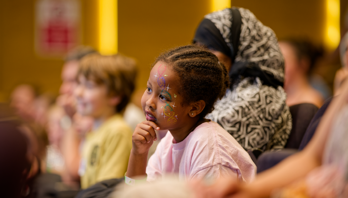 Young child with face paint attentively watching, surrounded by an audience in a warmly lit indoor setting.