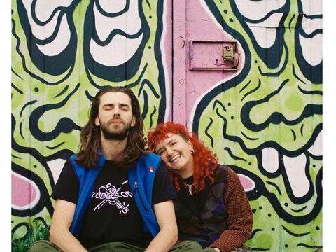 Two people sit on ground against brightly coloured cartoon spraypainted wall