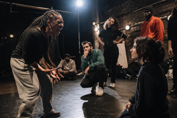 An individual passionately performs in a theater workshop, surrounded by attentive participants, under dramatic stage lighting.