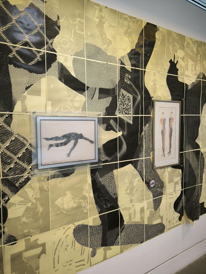 A gallery wall installation features a large yellow-and-black graphic mural of overlapping human silhouettes with two framed artworks mounted on top along a white corridor.
