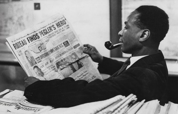 A man in a suit reads a newspaper while smoking a pipe, sitting at a table with more newspapers.