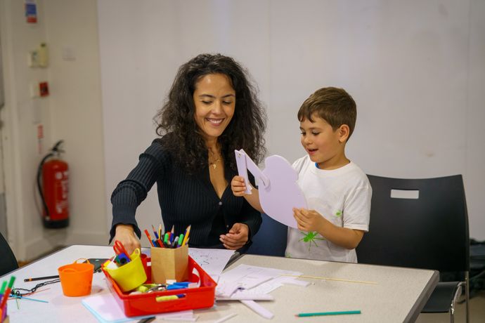 Woman and child happily crafting at a table with pens, paper, and art supplies. The child holds a paper cutout.