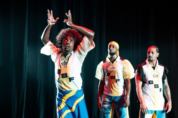 Three performers on stage in vibrant costumes, one gesturing expressively while the others stand attentively. Dark stage background.