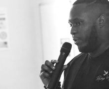 A man holding a microphone speaks, wearing a "Rising Hope" shirt, in a black and white setting.