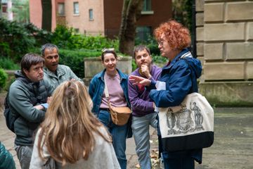 A group of people stands outdoors near a stone building and greenery, listening to a person speaking. The speaker wears a blue jacket and carries a white tote bag with a black classical illustration.
