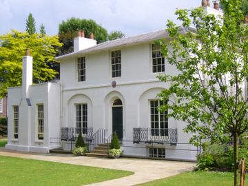 A two-story white Georgian-style house with large windows, surrounded by a green lawn and trees, under a partly cloudy sky.