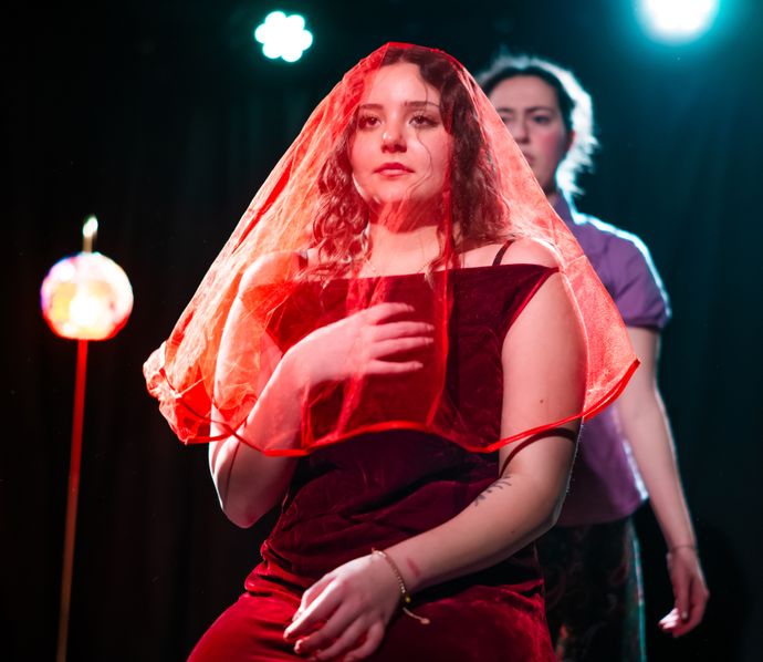 Young woman wearing red veil stands dramatically onstage with another person blurred behind under spotlights.