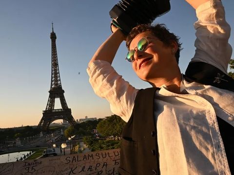 A person wearing sunglasses joyfully plays a concertina with the Eiffel Tower in the background, bathed in warm evening sunlight.