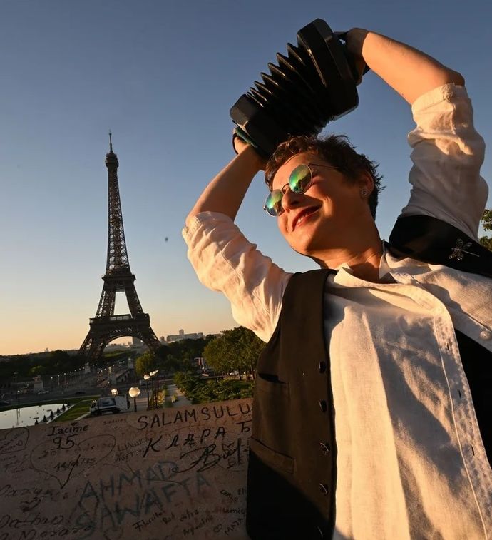 A person wearing sunglasses joyfully plays a concertina with the Eiffel Tower in the background, bathed in warm evening sunlight.