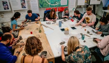 People seated around tables engaged in a pottery workshop, shaping clay with hands and tools, surrounded by pottery materials and informational posters.