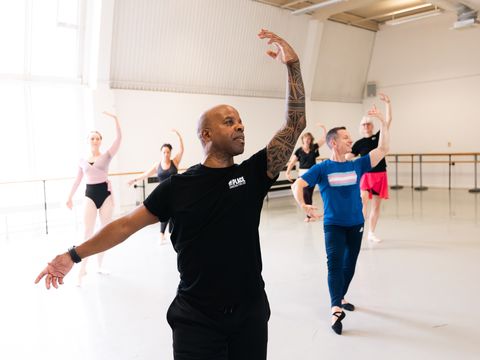 A ballet instructor leads a diverse group in a dance class. They are practicing arm positions in a spacious studio with large windows.