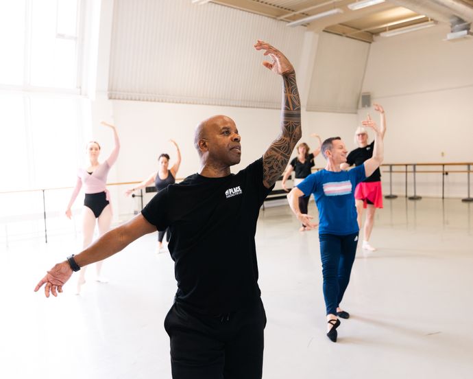 A ballet instructor leads a diverse group in a dance class. They are practicing arm positions in a spacious studio with large windows.