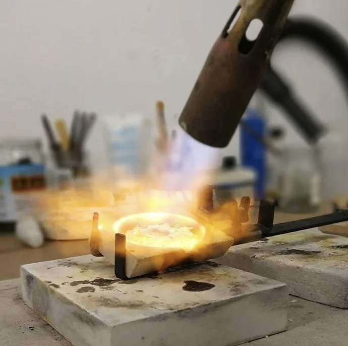 A blowtorch melting metal in a small crucible on a workbench, with tools and equipment in the blurred background.
