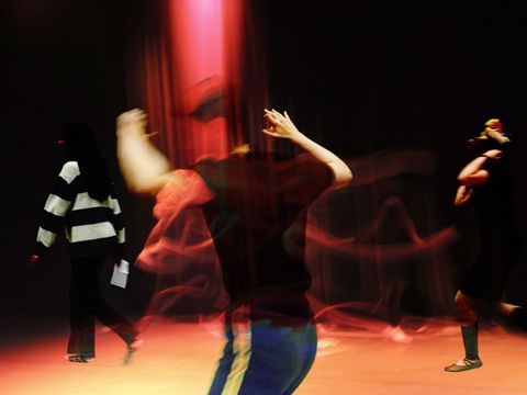 Blurred dancers in motion under red spotlight, one wearing a striped shirt, creating a dynamic and energetic scene against a dark background.