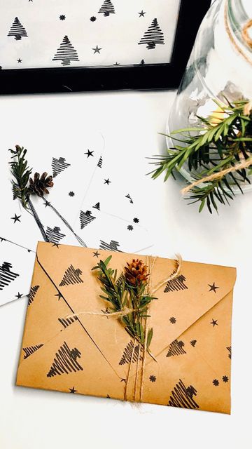 A rustic holiday-themed envelope wrapped in twine and decorated with pine sprigs and a small cone, surrounded by matching tree-patterned paper and a jar with greenery.