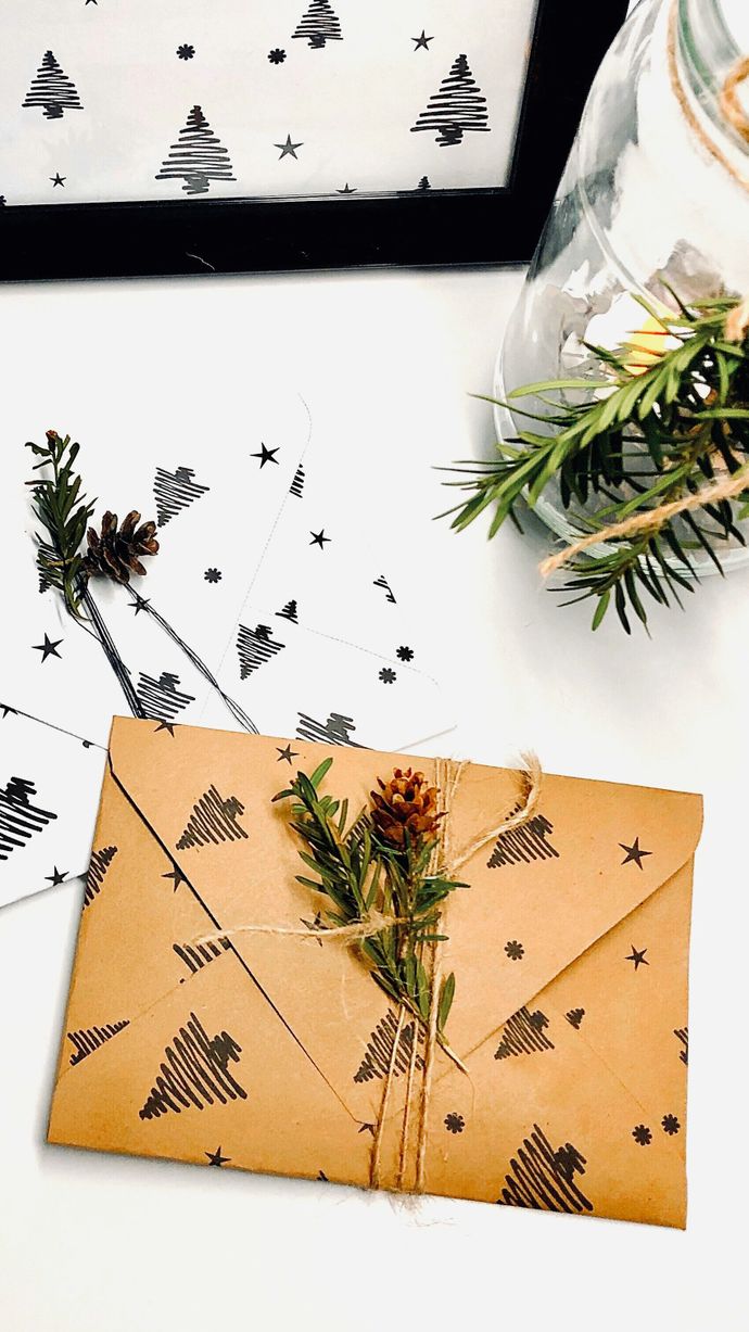 A rustic holiday-themed envelope wrapped in twine and decorated with pine sprigs and a small cone, surrounded by matching tree-patterned paper and a jar with greenery.