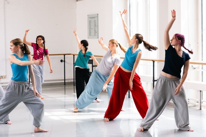 A group of dancers practice in a studio, wearing casual clothing, with arms raised, in natural light from large windows.