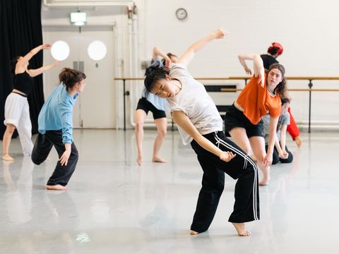 A group of dancers in varied movements practicing in a bright dance studio, wearing casual dance attire.