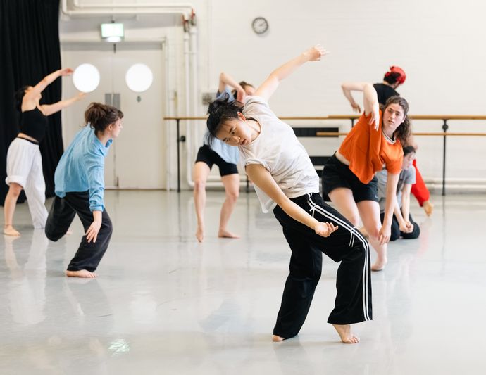 A group of dancers in varied movements practicing in a bright dance studio, wearing casual dance attire.