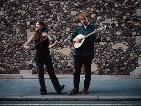 Two street musicians perform joyfully, playing violin and mandolin before a textured stone wall on a quiet street.