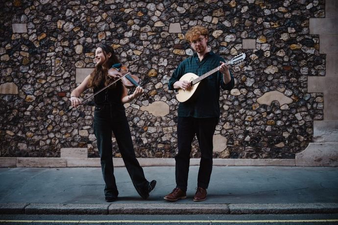 Two street musicians perform joyfully, playing violin and mandolin before a textured stone wall on a quiet street.