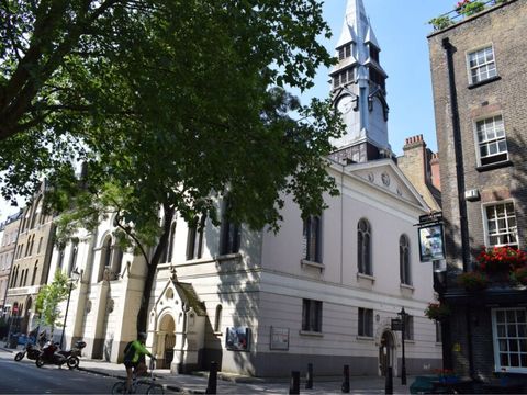 Street view of a historic church with a clock tower, leafy tree in foreground, and cyclist passing by. Nearby, a brick building with colorful flowers.