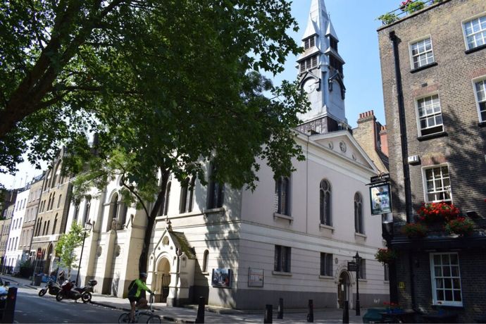 Street view of a historic church with a clock tower, leafy tree in foreground, and cyclist passing by. Nearby, a brick building with colorful flowers.