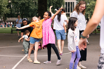Children and adults joyfully dancing and mingling in a park, with trees and buildings in the background.