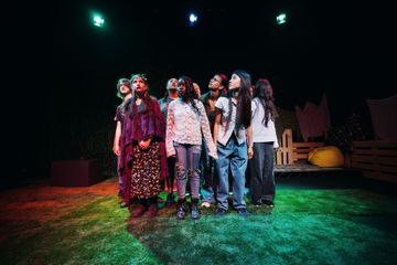 A group of people stands closely together on a stage, lit by colorful lights, with a simple set design in the background.