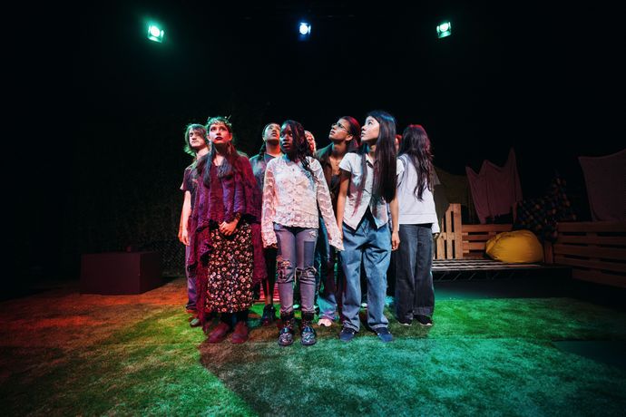 A group of people stands closely together on a stage, lit by colorful lights, with a simple set design in the background.
