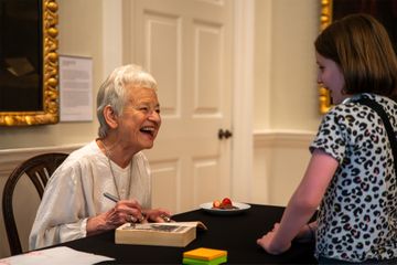 Jacqueline Wilson signing a book while smiling at a young child in a patterned shirt, inside a room with framed art and a plate of strawberries nearby.