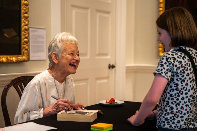 Jacqueline Wilson signing a book while smiling at a young child in a patterned shirt, inside a room with framed art and a plate of strawberries nearby.