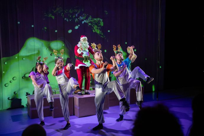 Four individuals dressed as reindeer standing in front of a person dressed as Santa on stage