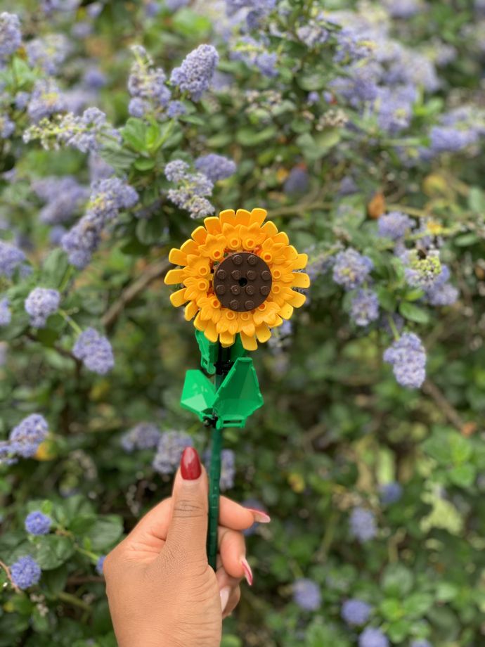 Hand holding a LEGO sunflower with a lush background of green leaves and clusters of light purple flowers.
