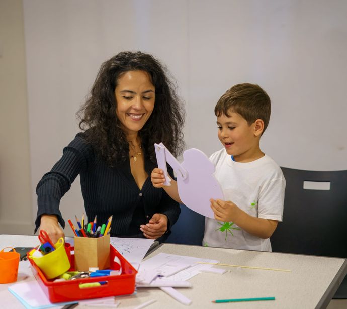 An adult and a child engaged in a craft activity at a table with art supplies, including markers, paper, and scissors.