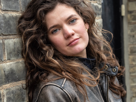 person with long brown curly hair looks straight to camera leaning against brick wall wearing brown leather jacket