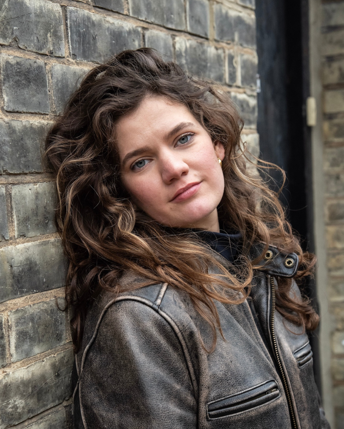 person with long brown curly hair looks straight to camera leaning against brick wall wearing brown leather jacket