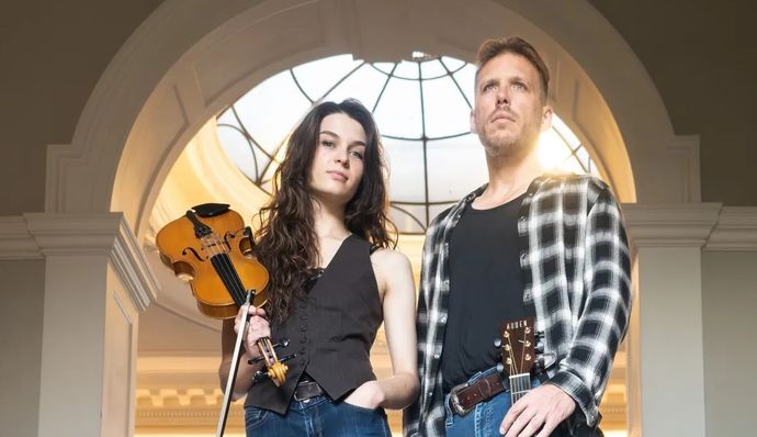 Two people stand under an arched window, one holding a violin and the other a guitar, dressed casually.
