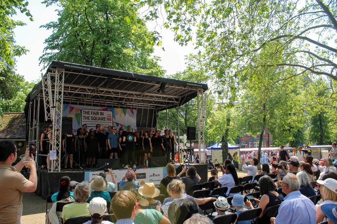 Outdoor performance on a stage with a choir and audience, surrounded by trees and sunlight at a fair.