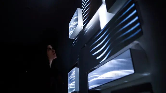 A person looks at a dark art installation with illuminated, wavy light patterns on protruding panels.