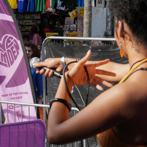 Performer with a microphone on stage, facing a crowd. A purple banner with a heart design is visible in the background.