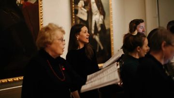 A choir group sings indoors, holding sheet music, with ornate paintings in the background.