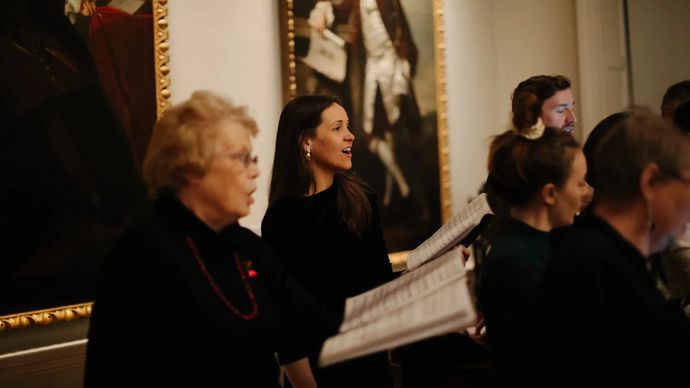 A choir group sings indoors, holding sheet music, with ornate paintings in the background.