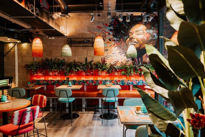 Vibrant restaurant interior with colorful mural, red and teal seating, pendant lights, and lush greenery along the walls.