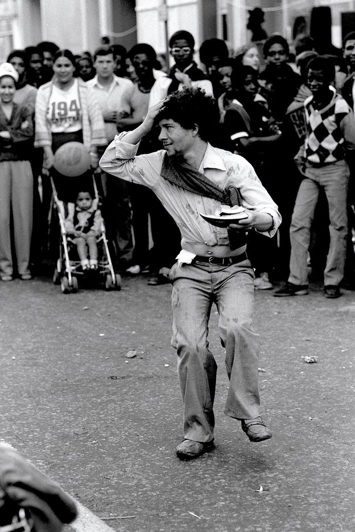Street performer juggling while balancing a plate on his head, surrounded by a crowd of onlookers, including a child in a stroller.