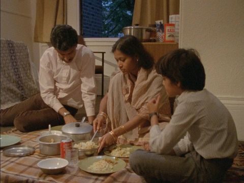 A family of three seated on the floor, sharing a meal, with various dishes and a soda can on a tablecloth in a cozy room.